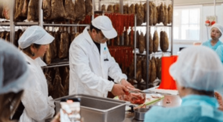 Workers in lab coats and caps handling meat in a processing facility.